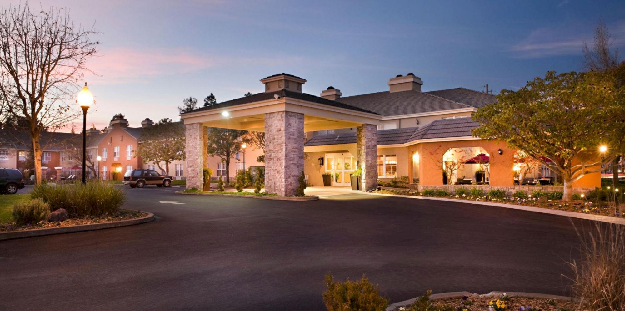 The image shows a well-lit, attractive building with a carport-style entrance, surrounded by trees and landscaped gardens, at twilight.