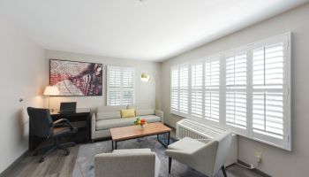 The image shows a bright living room with a couch, armchairs, a coffee table, a desk with a chair, and large windows with shutters.