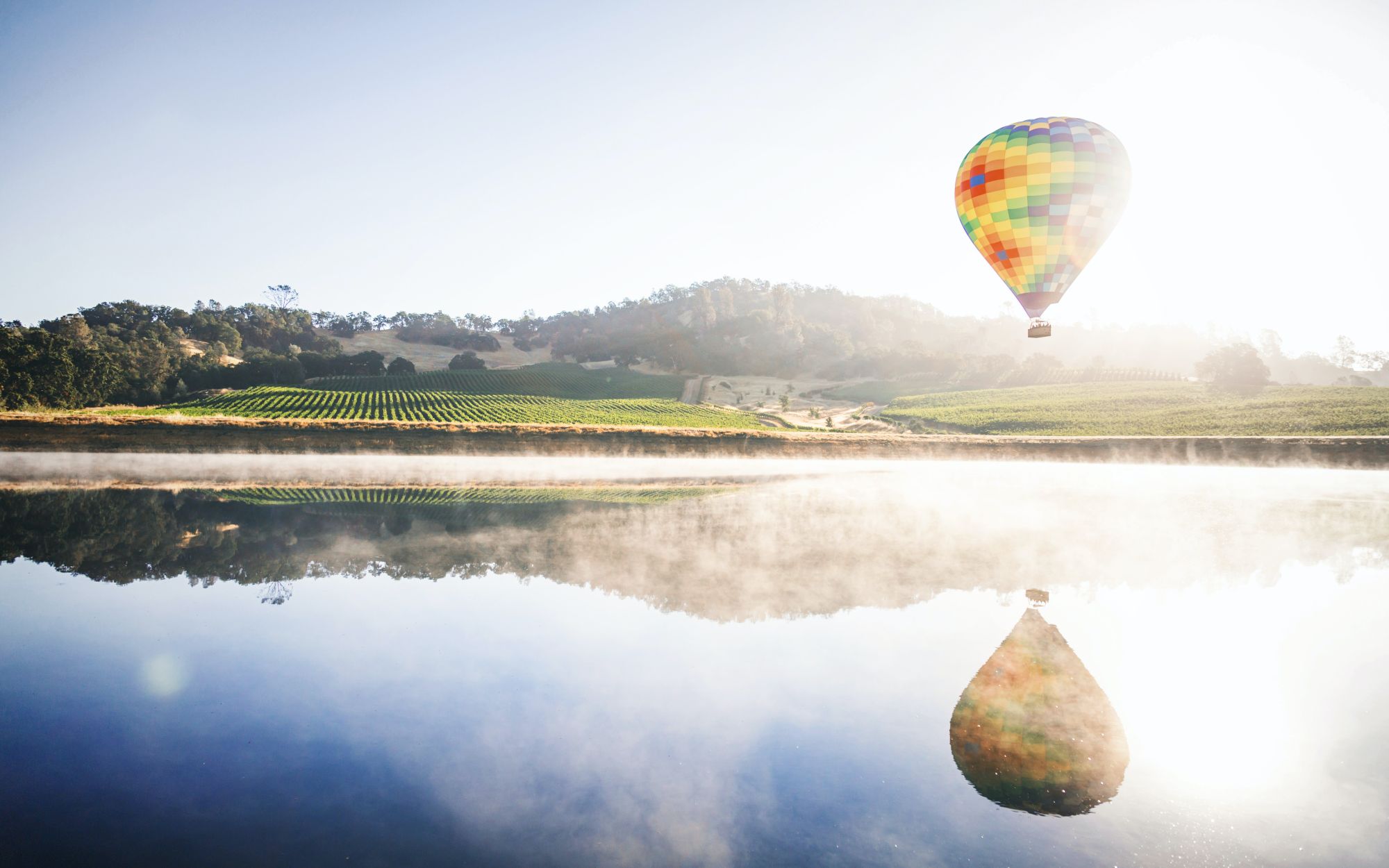 A colorful hot air balloon floats above a serene lake with hills and vineyards in the background, reflecting beautifully on the water's surface.