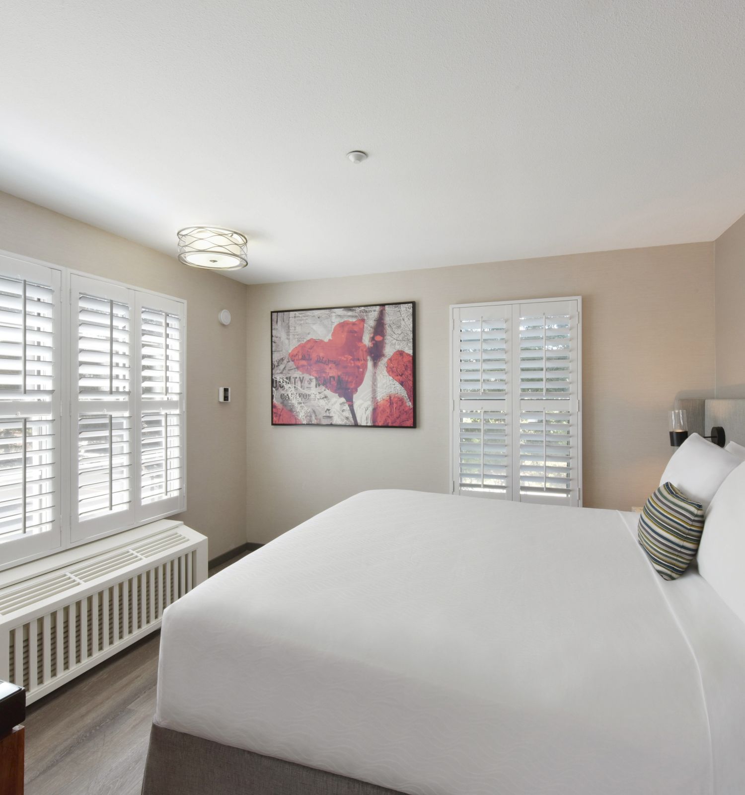 A modern, minimalistic bedroom with a large bed, two windows, white blinds, contemporary artwork, and a unique wall-mounted light.