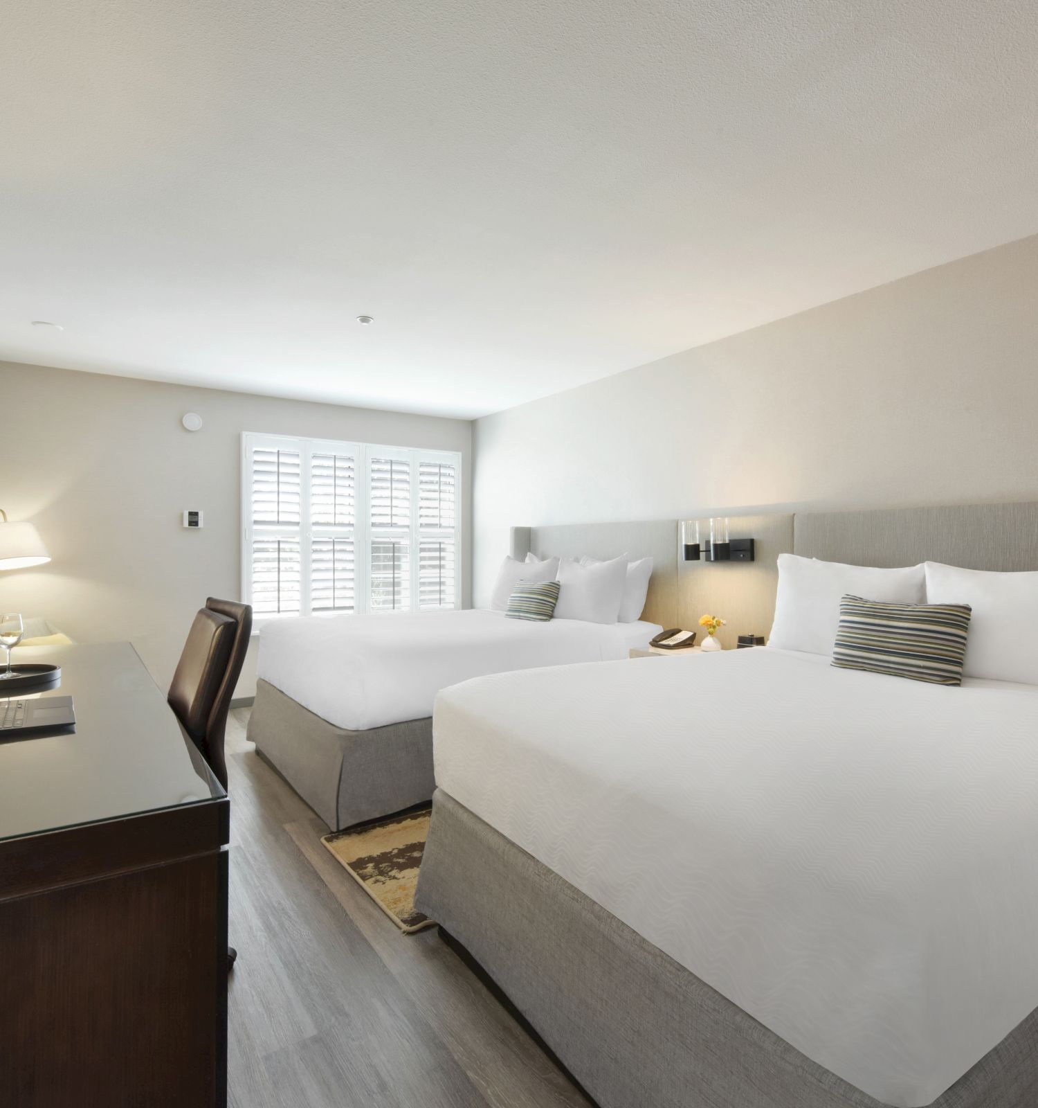 The image shows a hotel room with two large beds, a wooden desk, a chair, a television, a lamp, and a window with shutters.