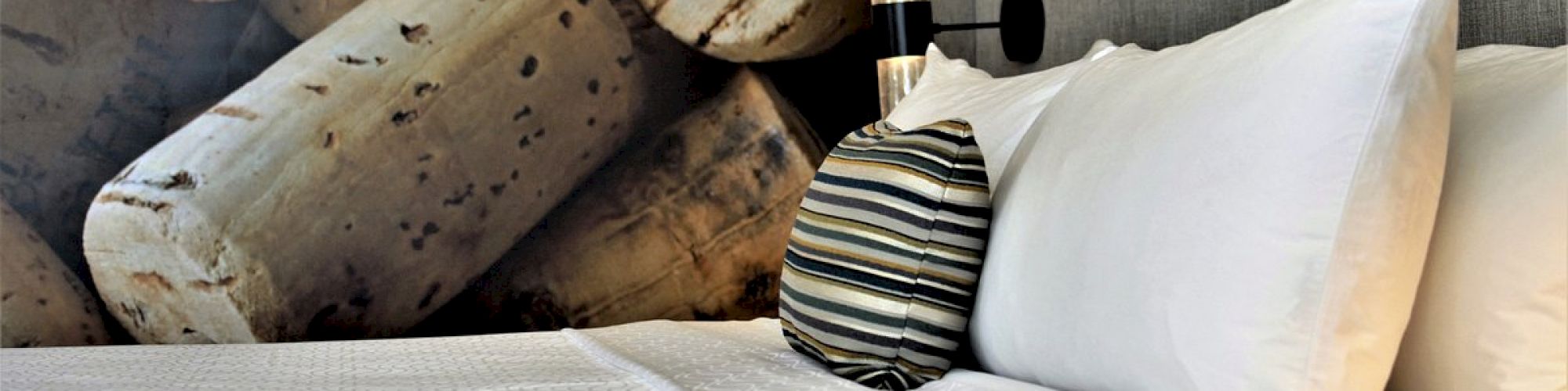 A neatly made bed with white linens and a striped pillow sits against a headboard with a large wall mural of corks in the background.