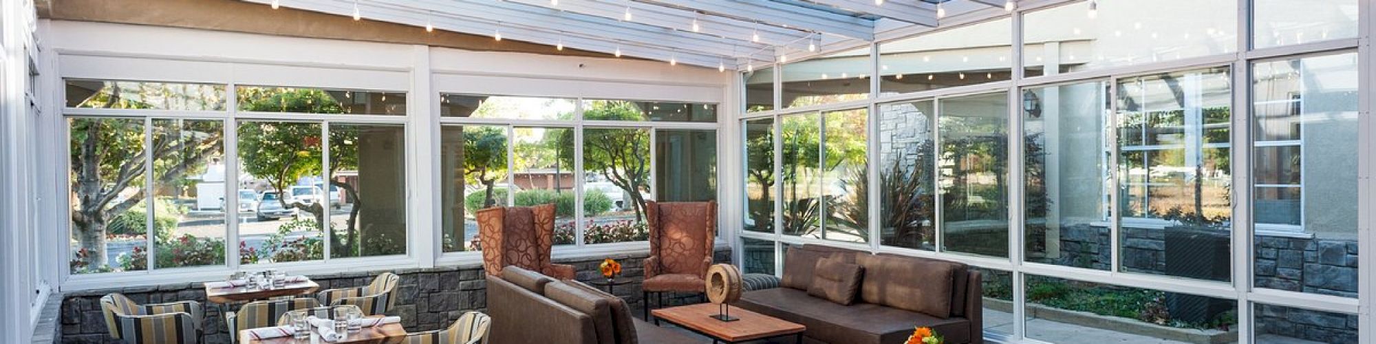 A spacious sunroom features a cozy seating area with sofas, armchairs, and tables. Large windows and a glass ceiling allow natural light to flood the space.