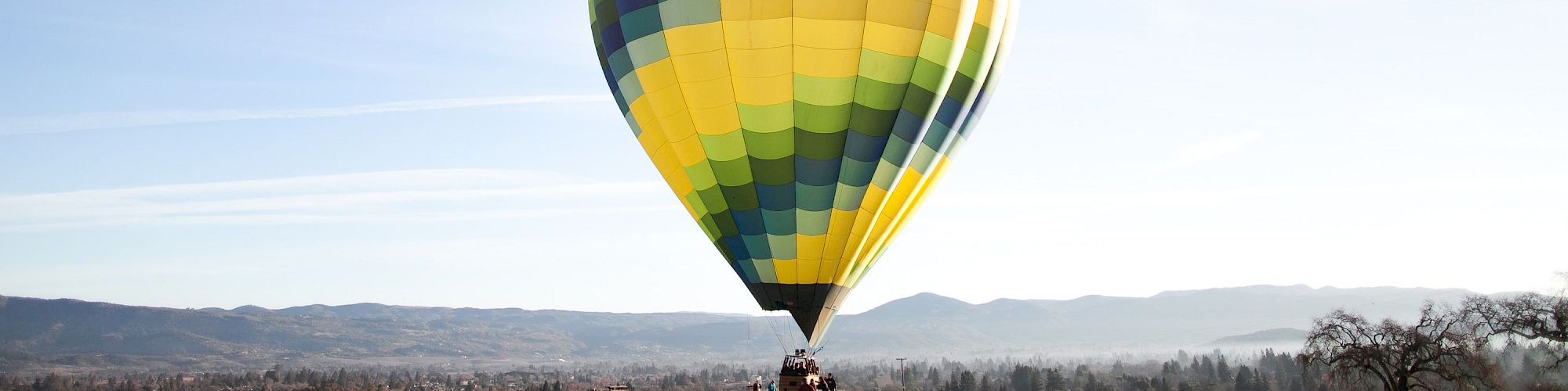 A large, colorful hot air balloon is about to take off from a field, with trees and mountains visible in the background.