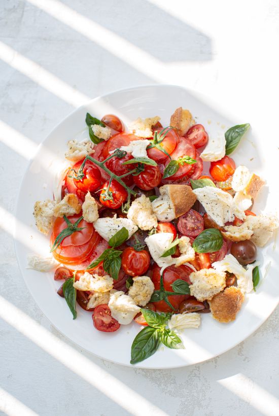 A fresh salad featuring tomatoes, mozzarella cheese, basil leaves, and croutons on a white plate, with sunlight streaming through blinds.