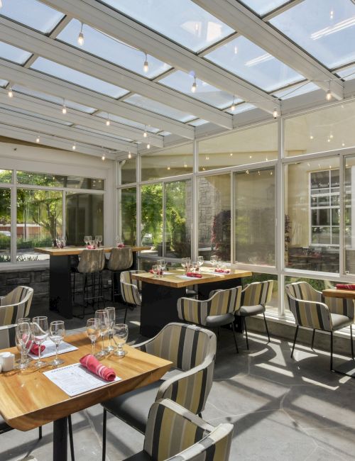 A modern, sunlit restaurant with glass walls and ceiling, featuring neatly set tables and striped chairs, ready for guests.