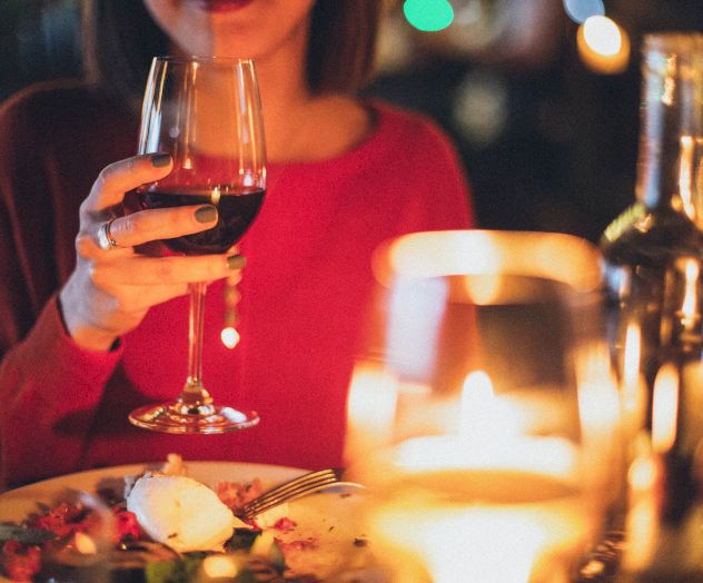 A person in a red outfit is holding a wine glass at a candlelit dinner table, with food and another bottle of wine.