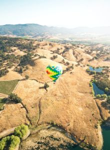 A colorful hot air balloon flies over a hilly, dry landscape with sparse vegetation, surrounded by small bodies of water and distant mountains.