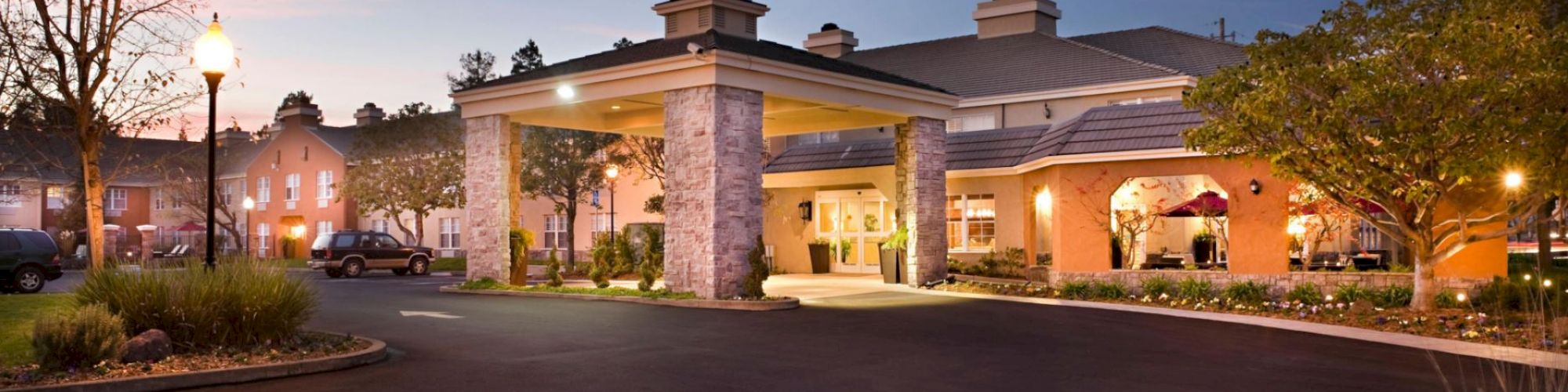 The image shows a large building with a welcoming entrance, possibly a hotel or residential facility, surrounded by landscaped grounds and illuminated lighting.