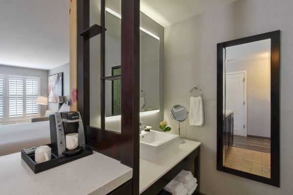 A modern hotel room with a coffee machine on a counter, a sink, a large mirror, and a visible bedroom area with a bed and a window in the background.