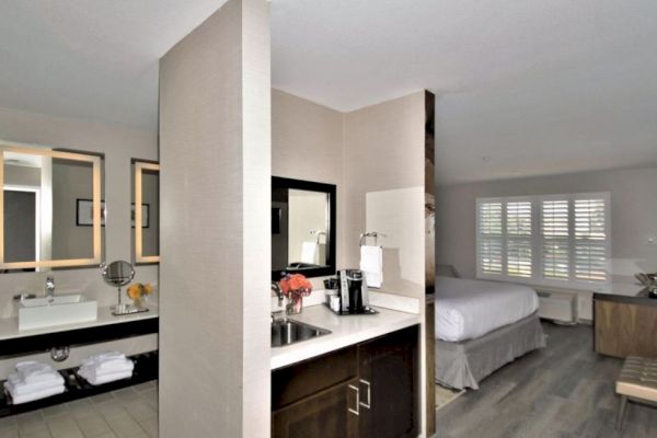 The image shows a modern hotel room with a bathroom area, a sink with mirrors, a counter with drawers, a bed, and a window with shutters.