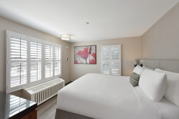 A well-lit bedroom with a large bed, white linens, window with blinds, a wall painting, and a small nightstand.