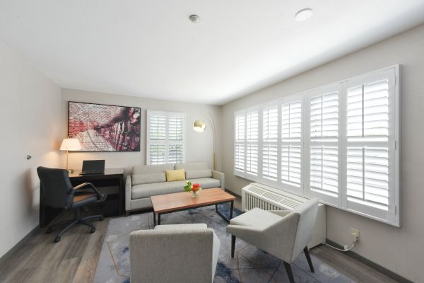 A bright living room with a seating area, a desk with a chair, a wall-mounted TV, large windows with shutters, and a small table with flowers.