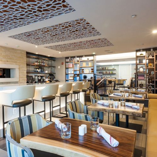 The image shows a modern restaurant interior with a bar area, stools, tables, chairs, napkin rolls, and shelves with various items.