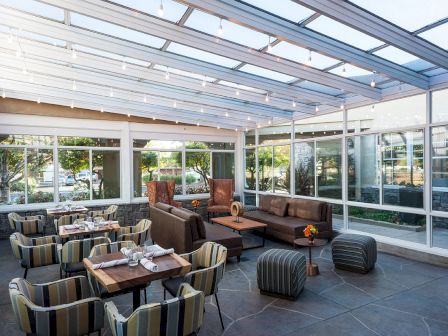 A modern, sunlit indoor seating area with cushioned chairs and sofas, large windows, and a glass ceiling, creating an inviting atmosphere for dining or relaxing.