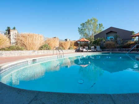 The image shows an outdoor swimming pool area with clear blue water, surrounded by lounge chairs, umbrellas, and a building in the background.