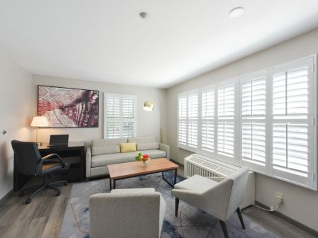 A bright living room features a couch, chairs, a coffee table with flowers, a desk with a computer, artwork, and large windows with shutters.