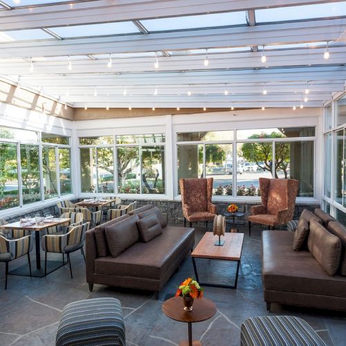 A modern, sunlit indoor seating area with comfortable couches, chairs, tables, and large windows providing a view of the outside.