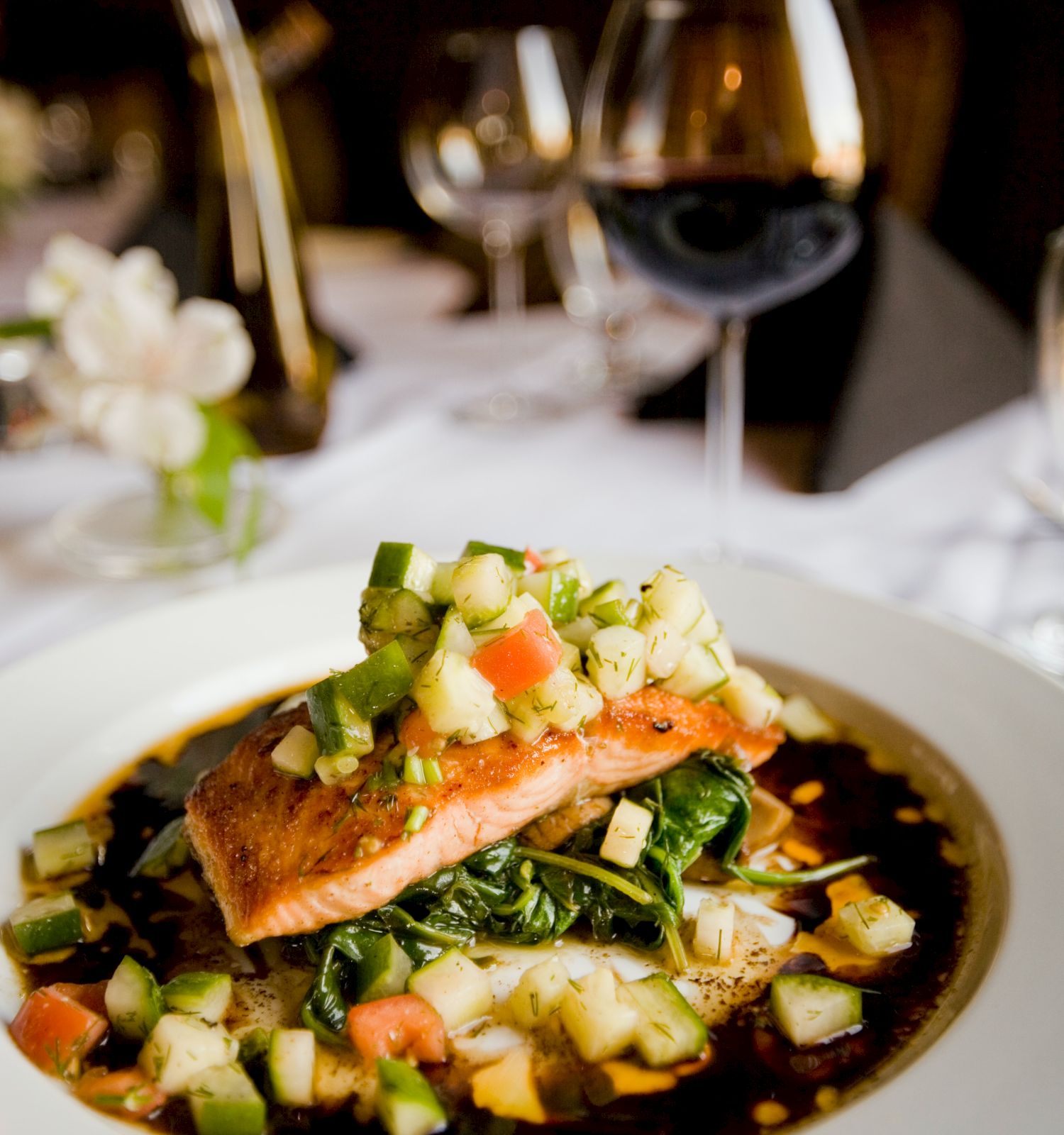 A plated dish featuring a piece of salmon topped with diced vegetables and served over sautéed greens, accompanied by a glass of red wine.