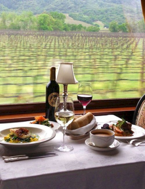 A dining setup in a train with wine glasses, a bottle, plated dishes, bread, and a window view of a vineyard outside.