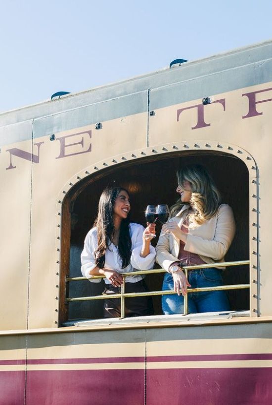 Two women are standing at the window of a train car, holding wine glasses and smiling at each other.