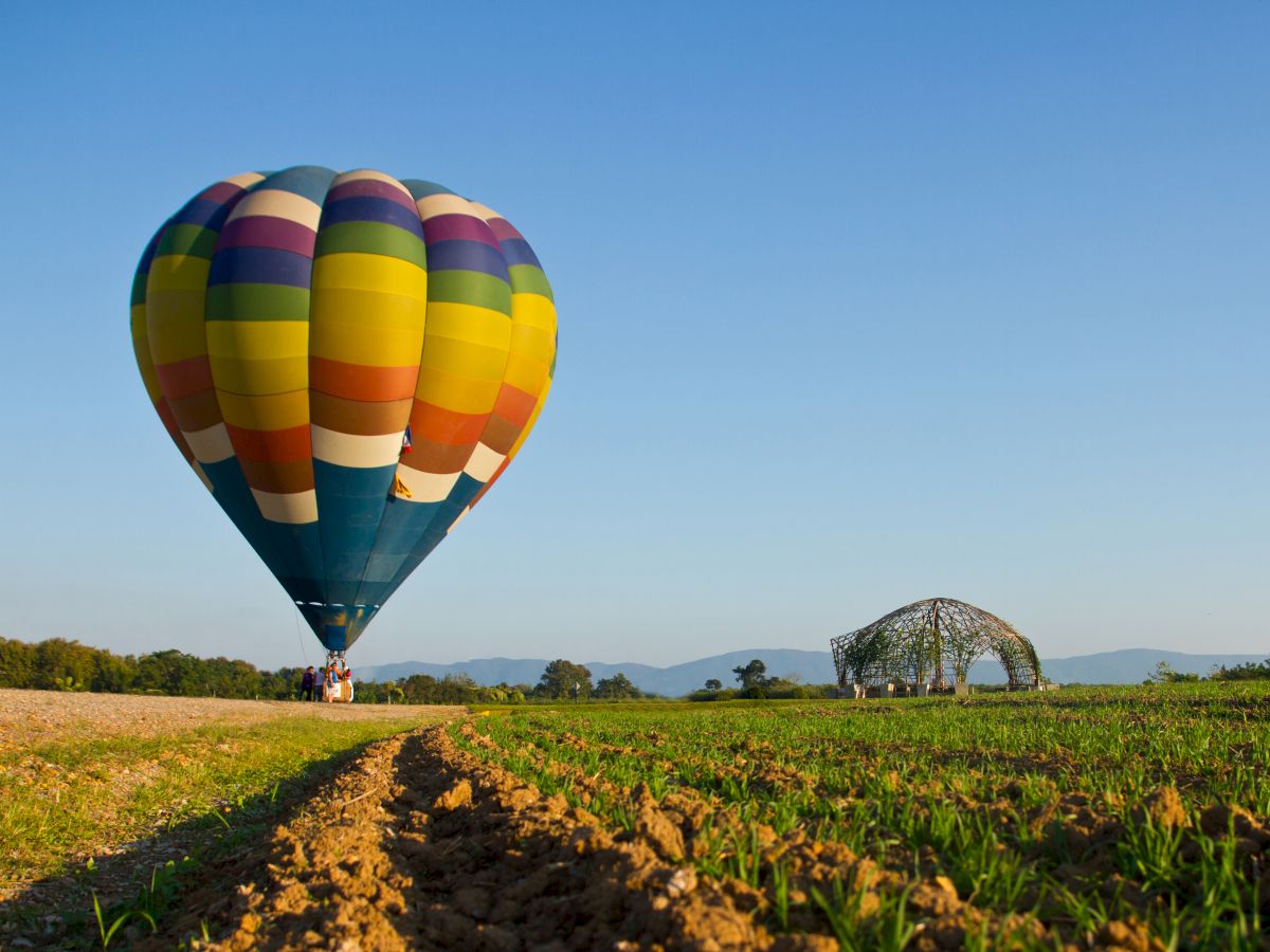 A colorful hot air balloon is taking off or landing in an open field with a clear blue sky, and a structure is visible in the background.