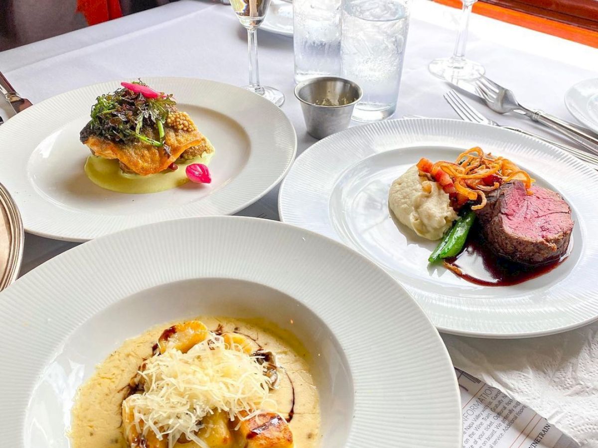 An elegantly set table with three gourmet dishes, two glasses of sparkling wine, and the hands of a person possibly browsing a menu, always ending the sentence.