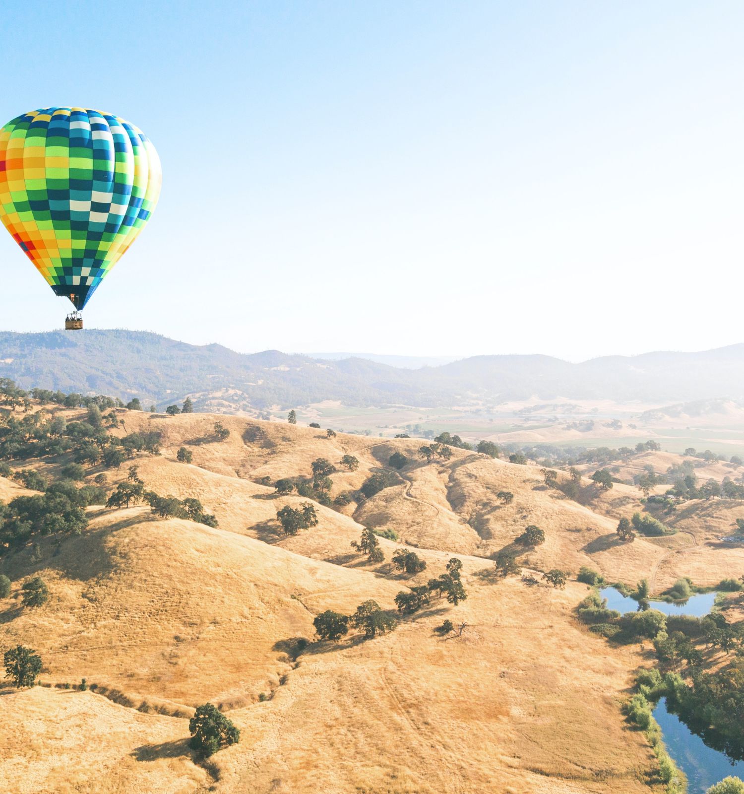 A colorful hot air balloon flies over a scenic landscape with rolling hills, patches of greenery, and a small body of water in the distance.