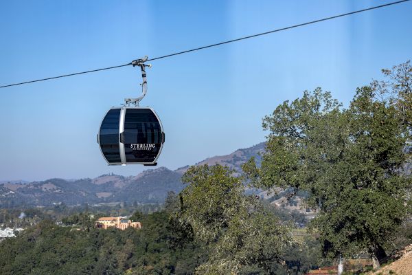 A gondola lift carries passengers over a scenic landscape with trees and distant hills under a clear sky, labeled 