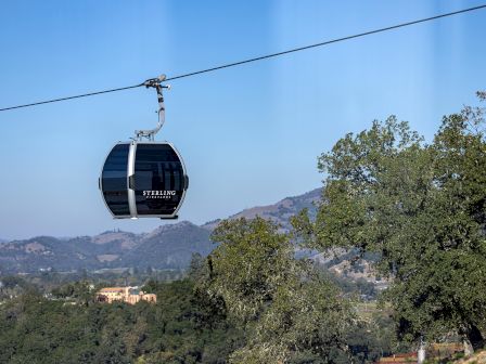 The image shows a cable car suspended in the air against a background of hills, trees, and a clear blue sky, with 